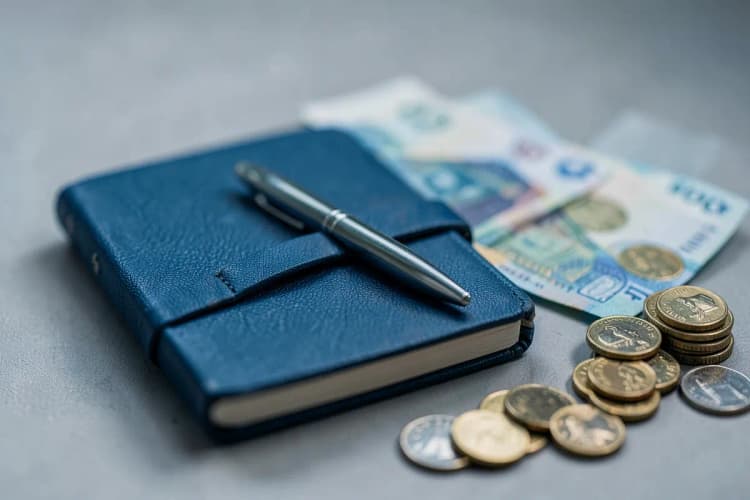 Uma pessoa anotando despesas em um pequeno caderno com uma caneta, ao lado de algumas moedas e notas.
